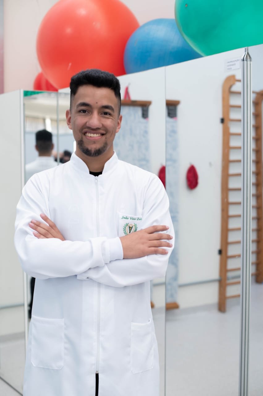 João Zvir em ambiente clínico, de braços cruzados e jaleco branco